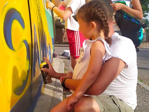 bambina che dipinge con gli spray su dei pannelli aiutata da suo padre