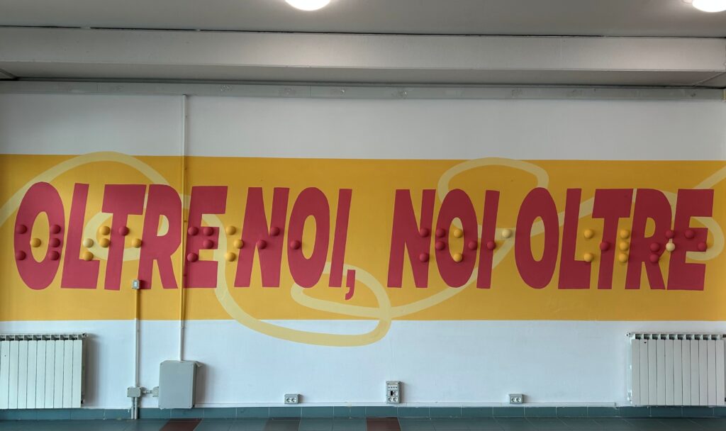 installazione artistica formata da una scritta con sopra delle semisfere in rilievo che riportano la stessa scritta "noi oltre, oltre noi" in scrittura braille, opera realizzata nell'istituto scuolastico piero sraffa di crema da goran castellotti